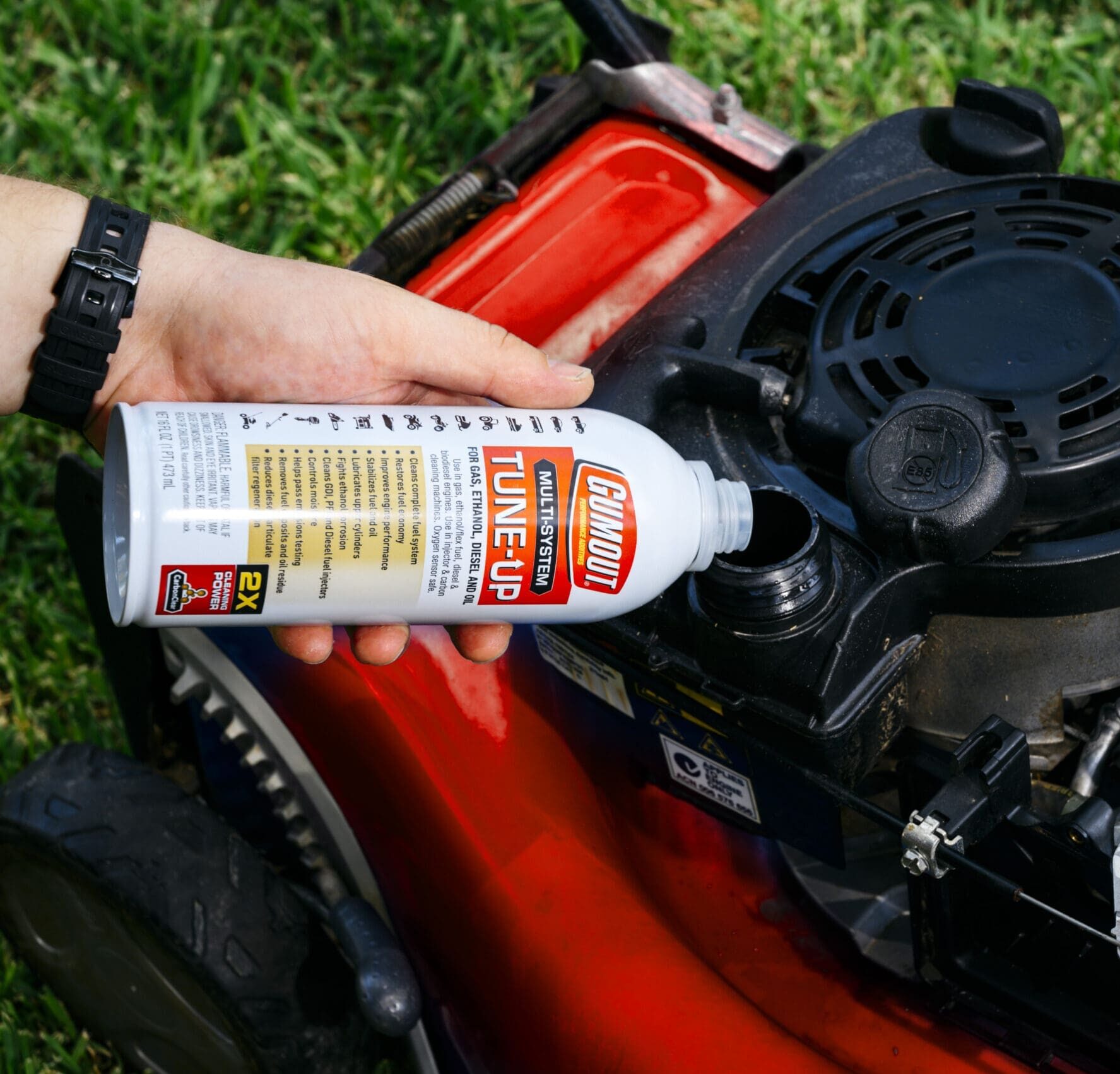 Person using an aerosol spray can to lubricate a small lawn mower engine, outdoors on grass.