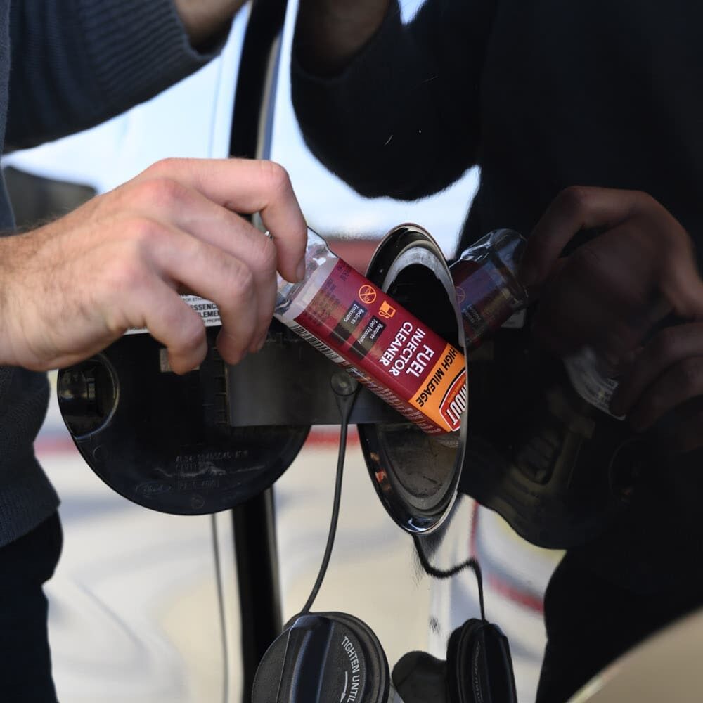 Close-up of hands pouring liquid from a red container into a car's open fuel tank inlet under a gas cap supervision by another person.