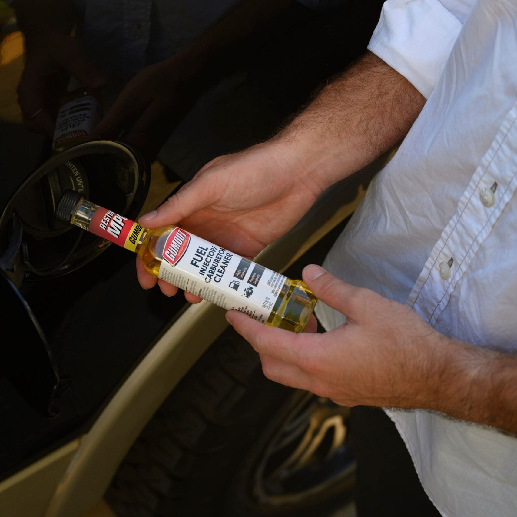 Person wearing a light-colored shirt holds a bottle of Gumout fuel injector cleaner near a car engine, ready to add the cleaner