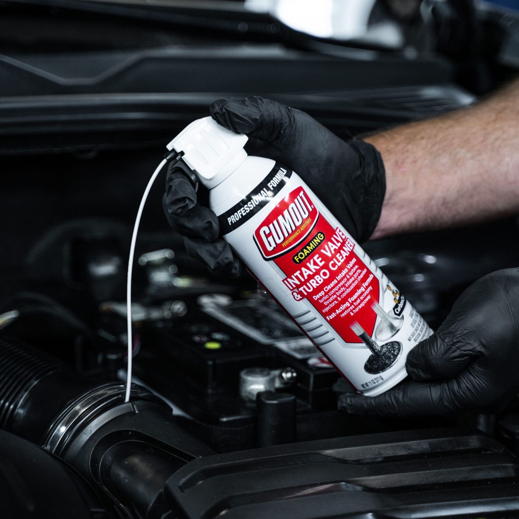 Gloved hands hold a Gumout intake valve cleaner spray can over a car engine bay, ready to spray cleaner into the engine section.